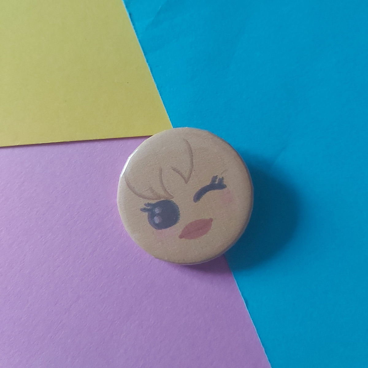 A round pinback button badge featuring a cute cartoon chick face with winking eyes, resting on a colourful background of blue, yellow, and pink urple paper.