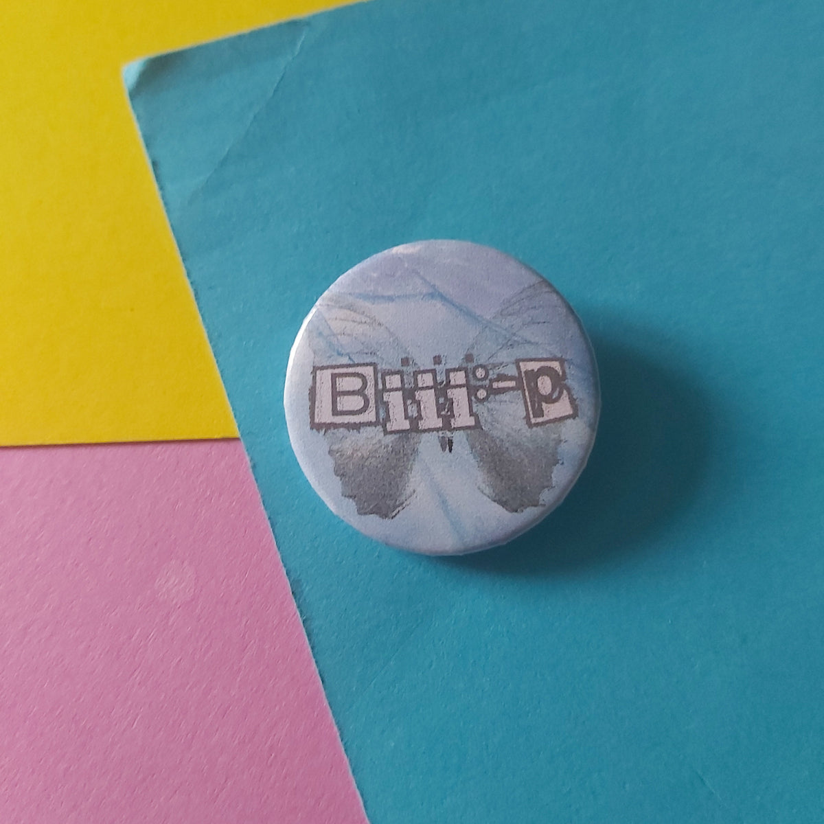 A round pinback button badge featuring the text 'Biii:-P' in a silver-grey outlined font over a silver butterfly with a light blue and white marbled background, placed on a colourful background of teal, yellow, and pink paper.
