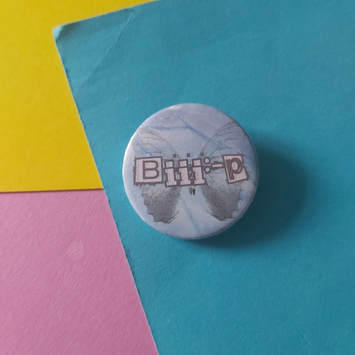 A round pinback button badge featuring the text 'Biii:-P' in a silver-grey outlined font over a silver butterfly with a light blue and white marbled background, placed on a colourful background of teal, yellow, and pink paper.