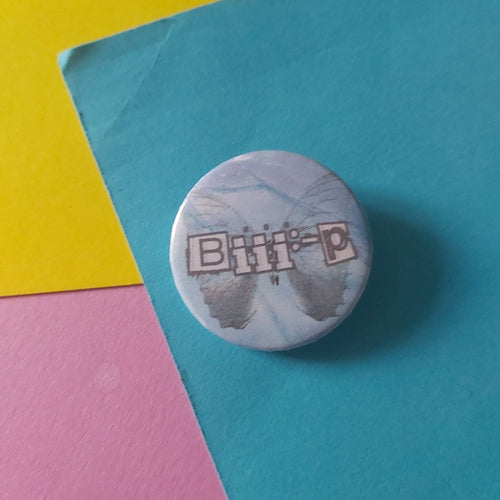 A round button pin with a white and blue marble pattern and the text 'Biii:-P' in a grey outlined box, placed on a colourful background of teal, yellow, and pink paper.