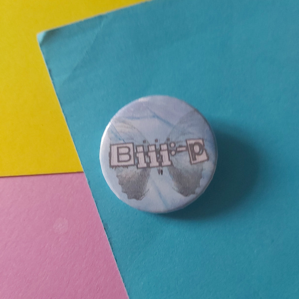 A round pinback button featuring the text 'Biii:-P' in a stylised font over a light blue and white marbled background, resting on a colorful background of teal, yellow, and pink paper.