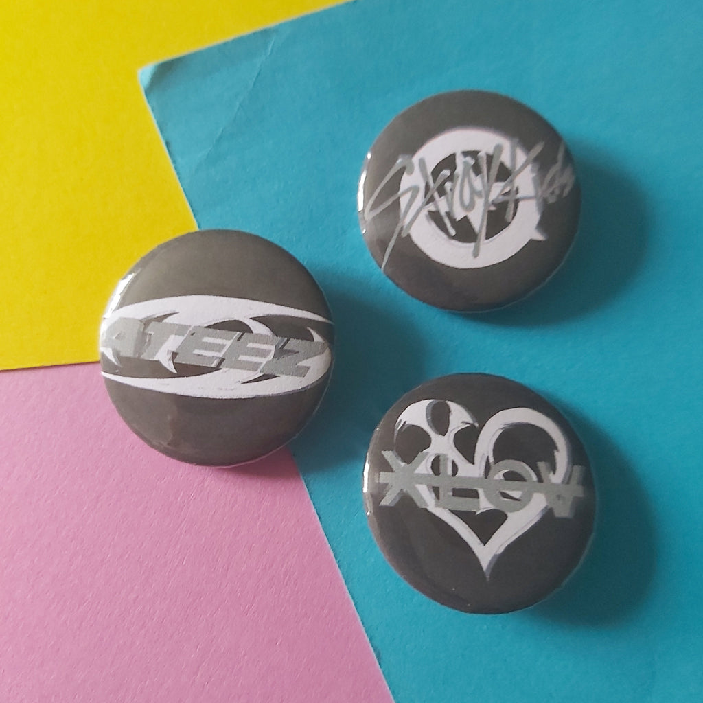 Three round black button badges with white graphic designs of K-pop logos, arranged on a colourful background split diagonally into yellow, blue, and pink sections.