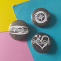 Three round black button badges with white graphic designs of K-pop logos, arranged on a colourful background split diagonally into yellow, blue, and pink sections.