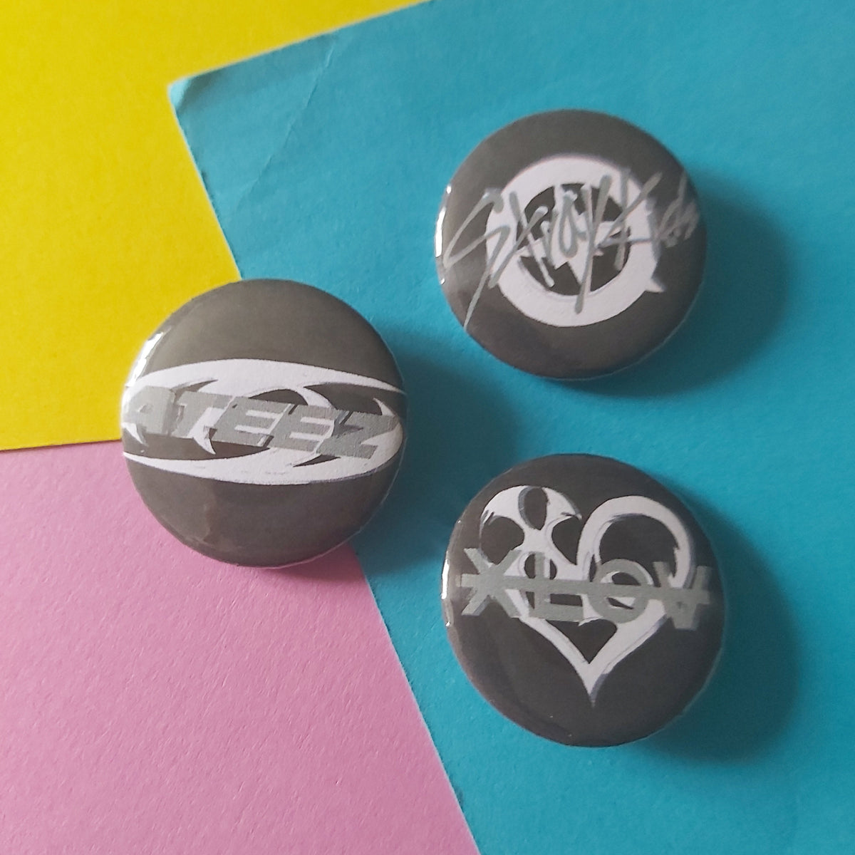Three round black button badges with white graphic designs of K-pop logos, arranged on a colourful background split diagonally into yellow, blue, and pink sections.