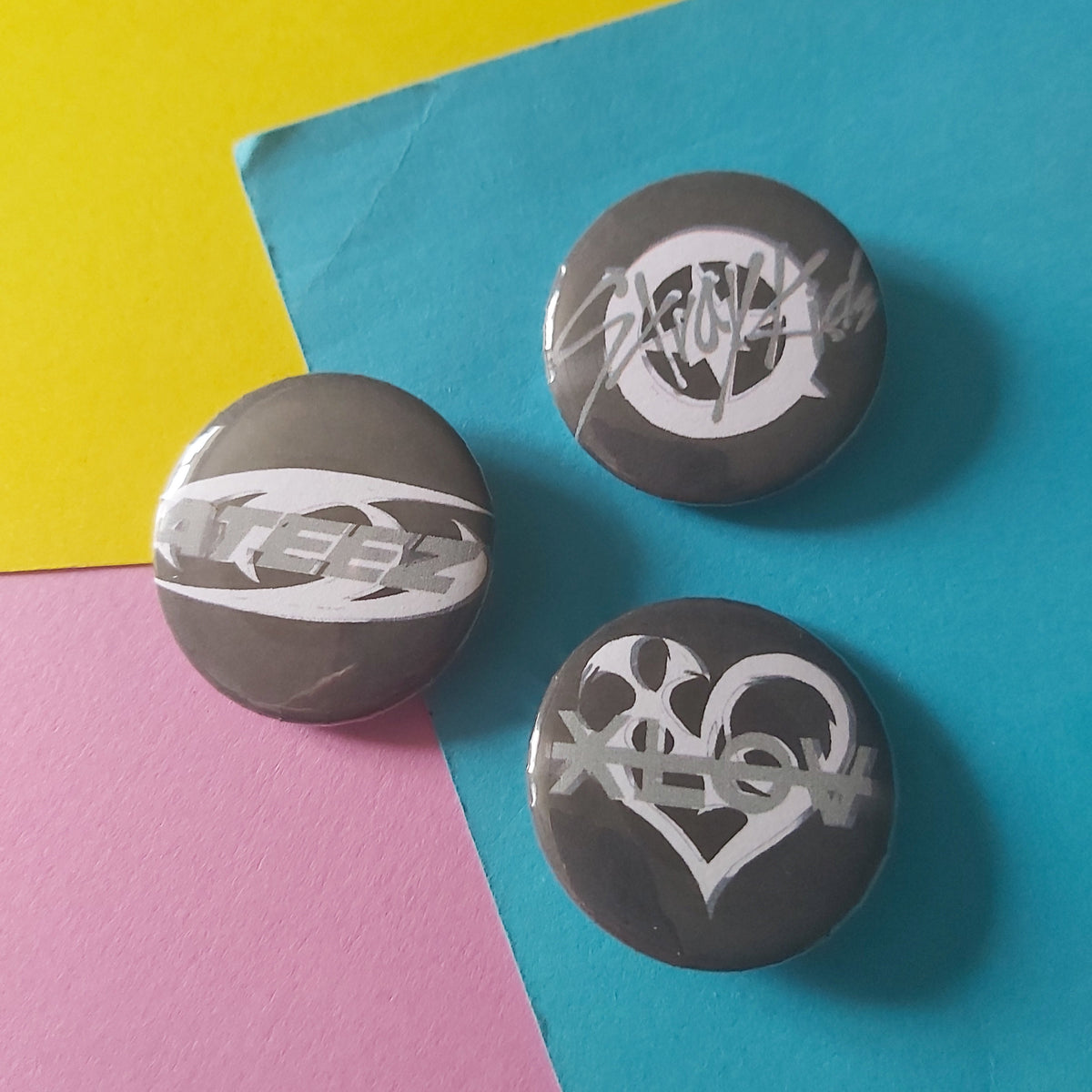 Three round gray pinback buttons with white stylized band logos, arranged on a colorful background of yellow, blue, and pink paper.