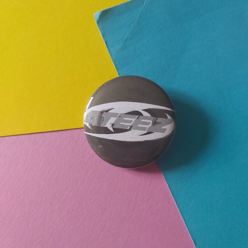 A small round product with a dark grey and white graphic design featuring the word 'ATEEZ' lies on a background of three colored paper sheets: blue, yellow, and pink.