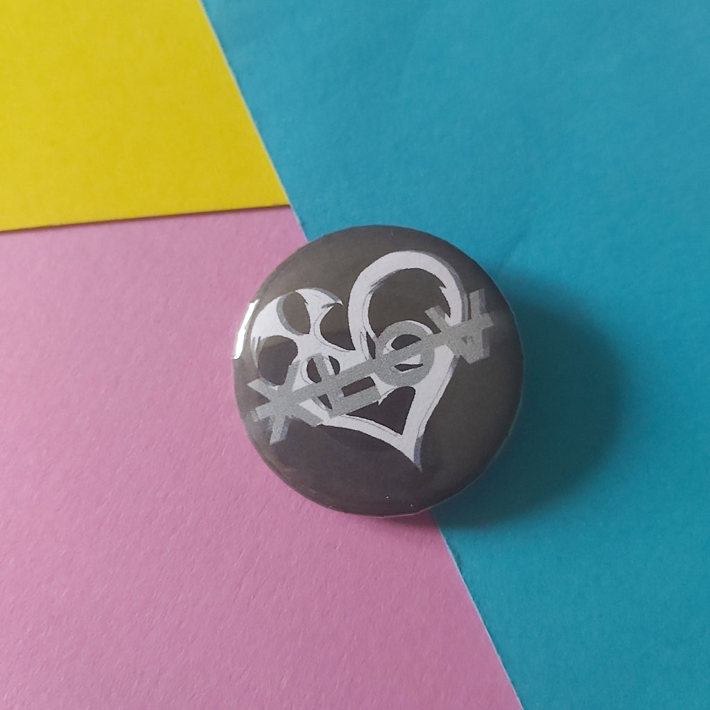 A round pinback button featuring a white stylised graphic of the Xlov group on a dark grey background, set against a colourful background of blue, pink, and yellow paper.