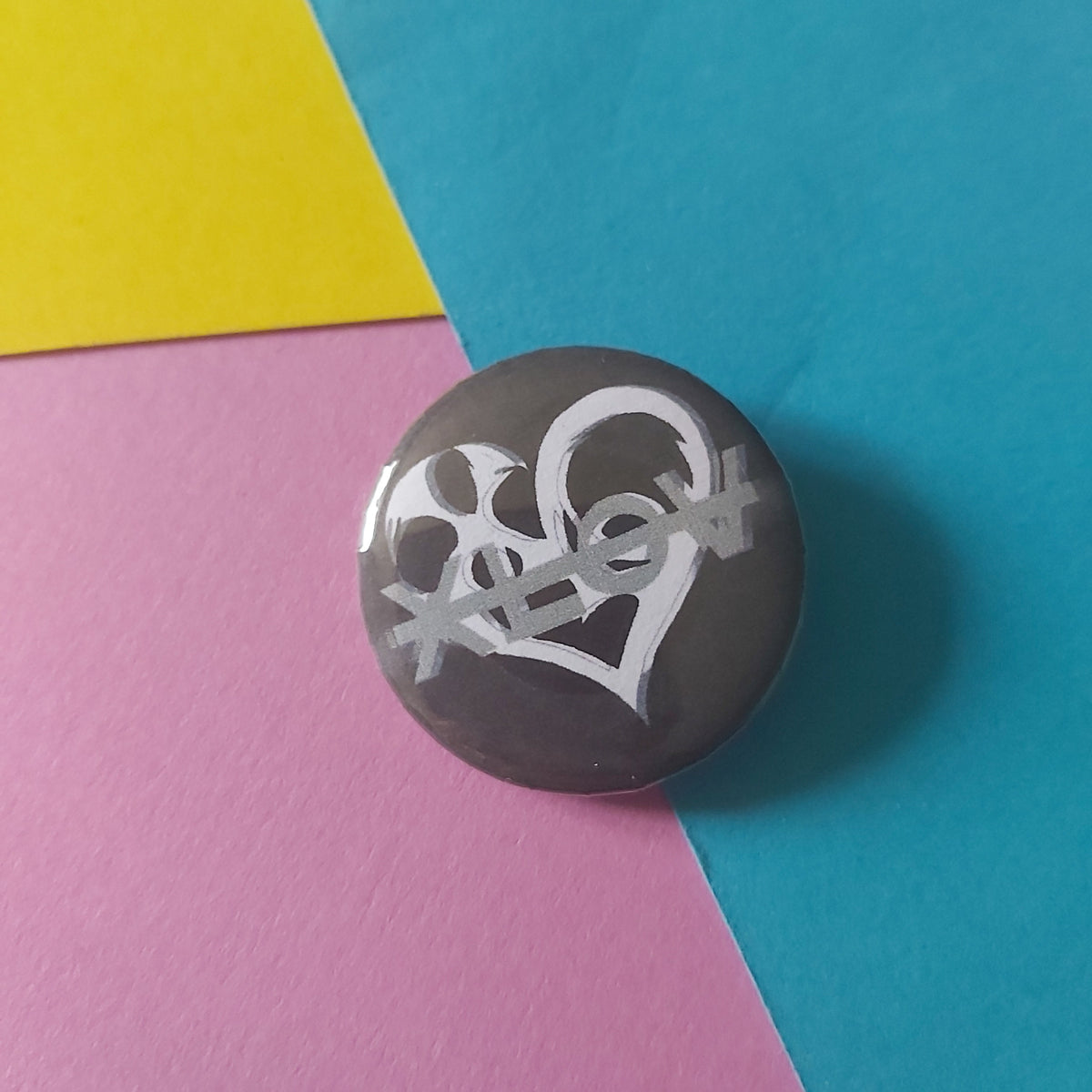 A round pinback button featuring a white stylised graphic of the Xlov group on a dark grey background, set against a colourful background of blue, pink, and yellow paper.
