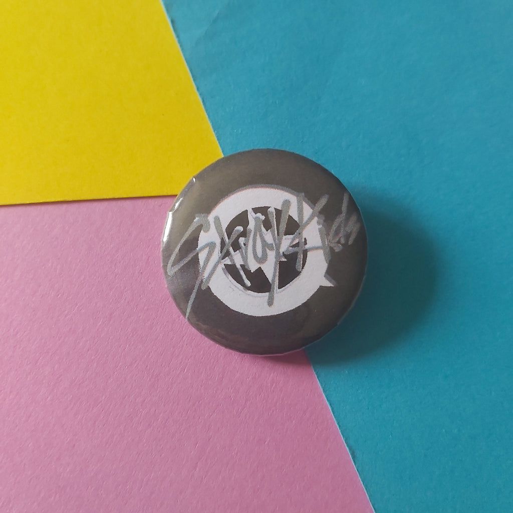 A round pinback button badge featuring a black-and-white Stray Kids compass logo and text design, resting on a diagonally split background of blue, pink, and yellow paper.