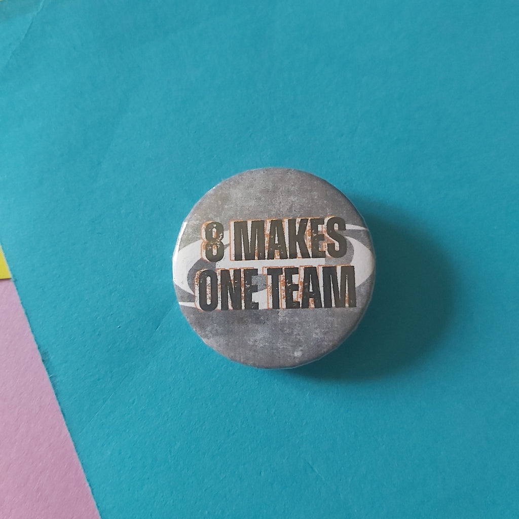 A round button badge with the text '8 MAKES ONE TEAM' with an orange outline in black on a grey and white Ateez logo background, placed on a bright blue surface with yellow and pink paper corners visible.