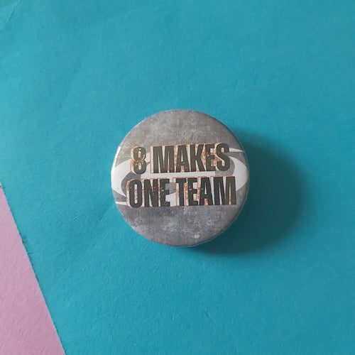 A round button badge with the text '8 MAKES ONE TEAM', placed on a bright teal background with small yellow and pink paper corners visible.
