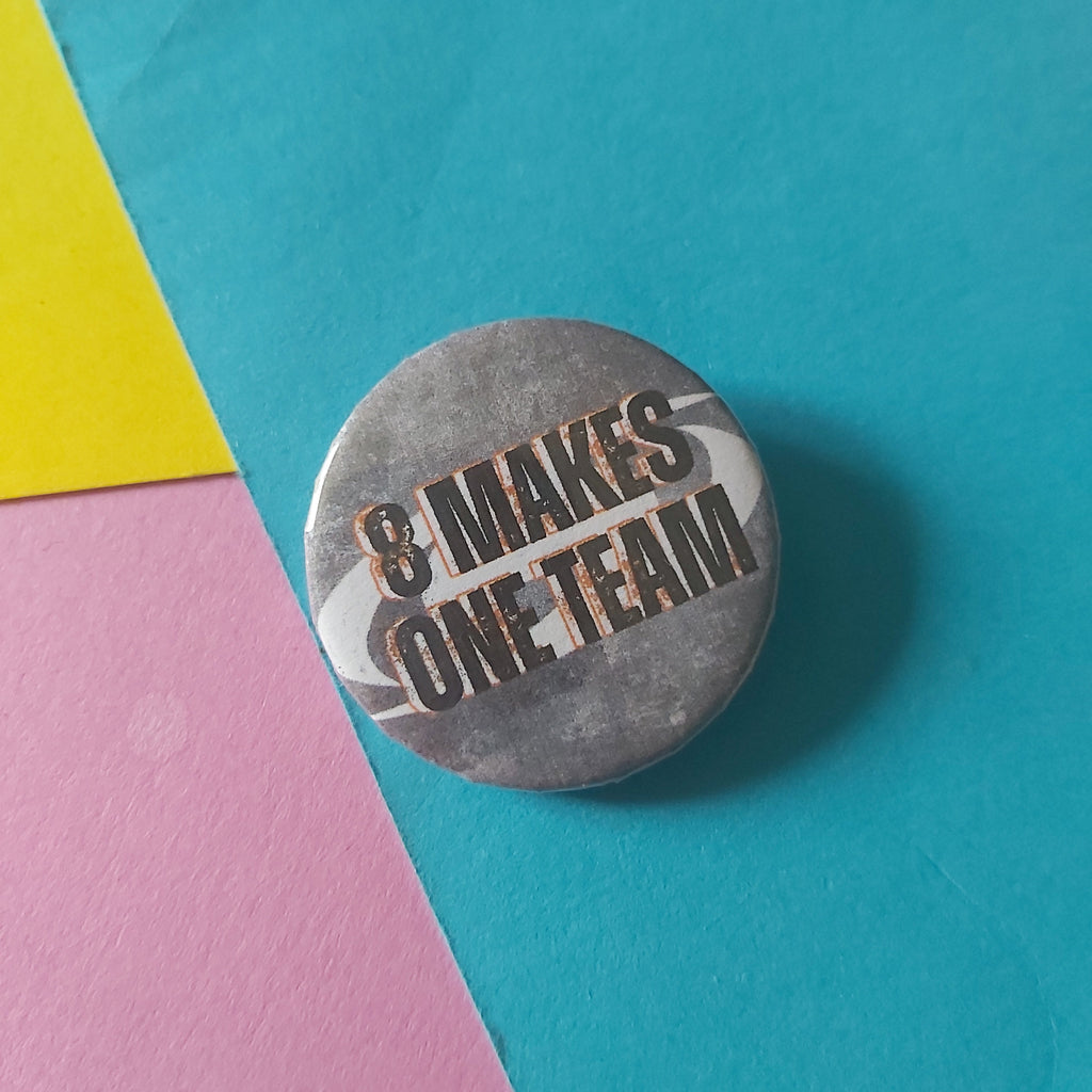 A round metal pin-back button badge with the text 'EIGHT MAKES ONE TEAM' in black and white, placed on a background of colored paper in teal, pink, yellow, and white.