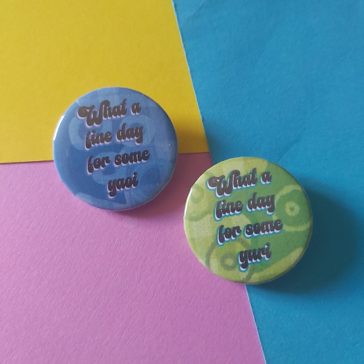 Two round pin-back buttons with the phrase 'What a fine day for some yuri' on a green background and 'What a fine day for some yaoi' on a blue background displayed on a colourful background of yellow, pink, and blue paper.