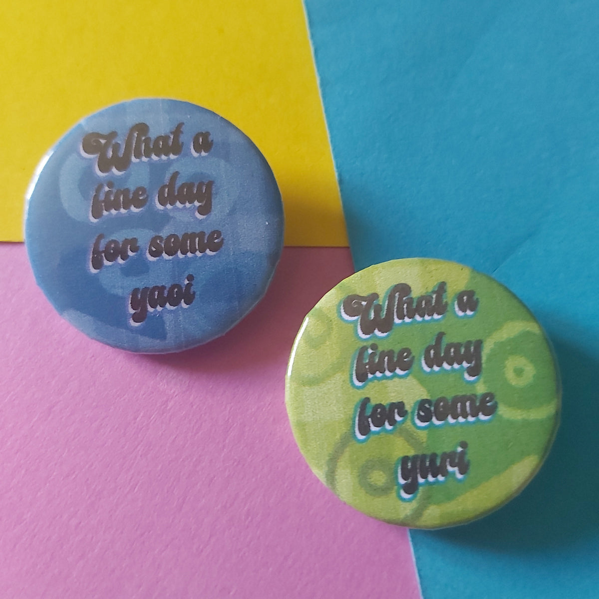 Two round pin-back buttons with the phrase ''What a fine day for some yuri' on a green background and 'What a fine day for some yaoi' on a blue background, both in white script, displayed on a split yellow, pink, and blue surface.