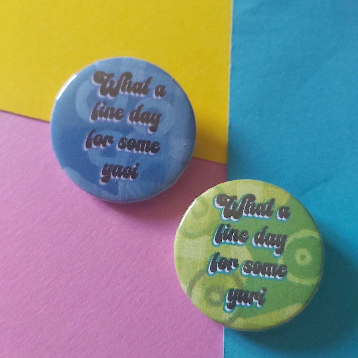Two round pin-back buttons with the phrase ''What a fine day for some yuri' on a green background and 'What a fine day for some yaoi' on a blue background, both in white script displayed on a colourful background of yellow, blue, and pink paper.