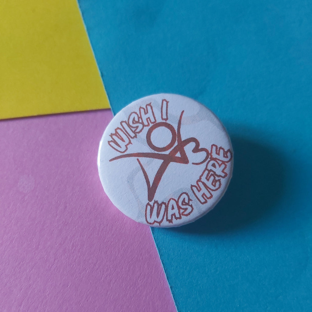 A round white button pin with a red graphic and text reading 'I Wish I Was Here' resting on a diagonally split background of blue, pink, and yellow paper.