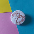 A round white button pin with a red graphic and text reading 'I Wish I Was Here' resting on a diagonally split background of blue, pink, and yellow paper.
