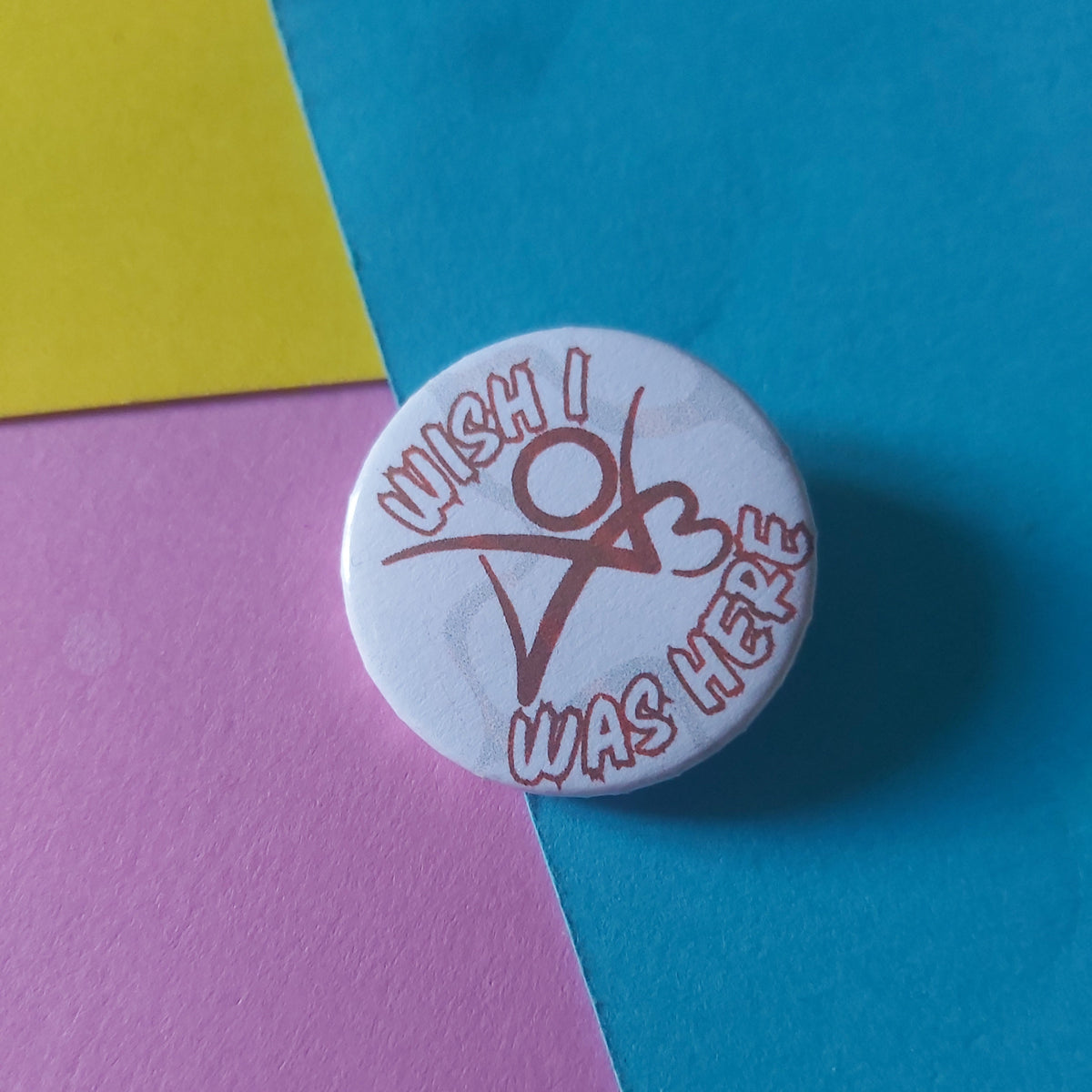 A round white button pin with a red graphic and text reading 'I Wish I Was Here' resting on a diagonally split background of blue, pink, and yellow paper.