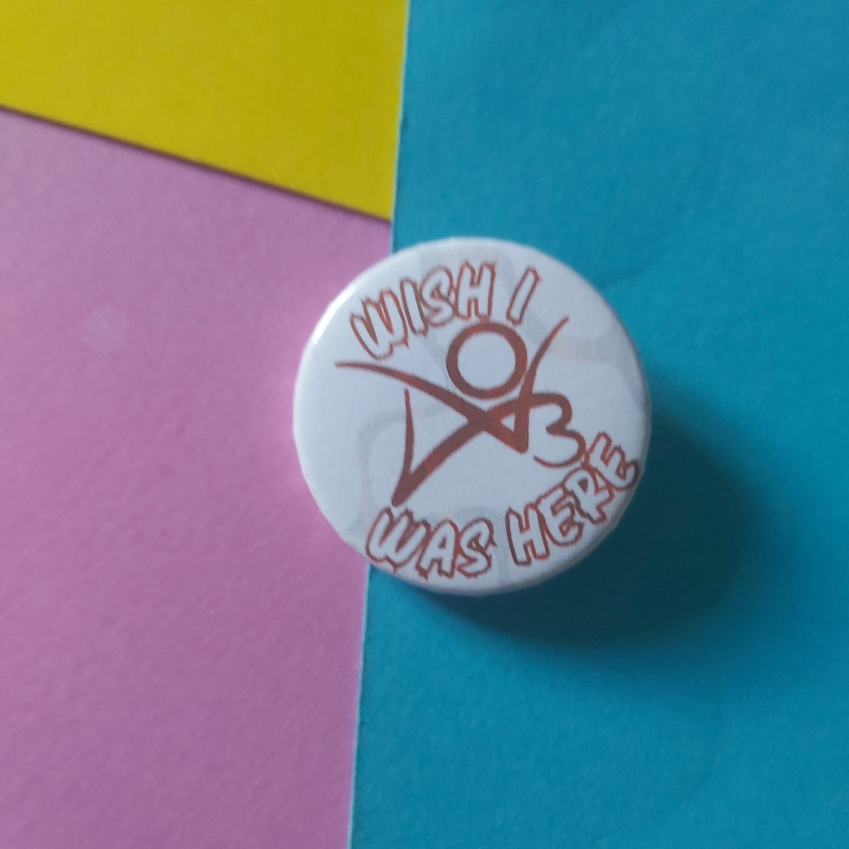 A round white button pin with a red graphic and text reading 'WISH I WAS HERE', placed on a colourful background of blue, yellow, and pink paper.