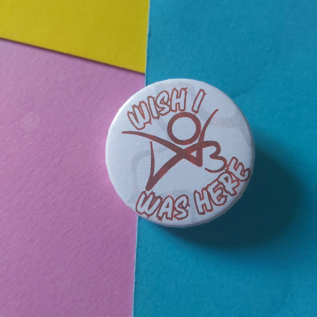 A round white button pin with a red graphic and the text 'WISH I WAS HERE', shown on a background split between blue, yellow, and pink paper.