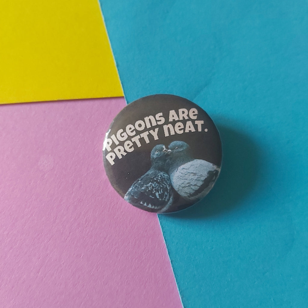 A round pinback button badge featuring a black background with two pigeons and the text Pigeons are pretty neat in a bold white font, placed on a colourful background of cyan, pink, and yellow paper.