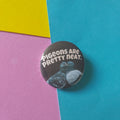 A round pinback button badge featuring a black background with white text reading 'Pigeons are pretty neat.' and grey pigeons, placed on a colourful background of teal, pink, and yellow paper.