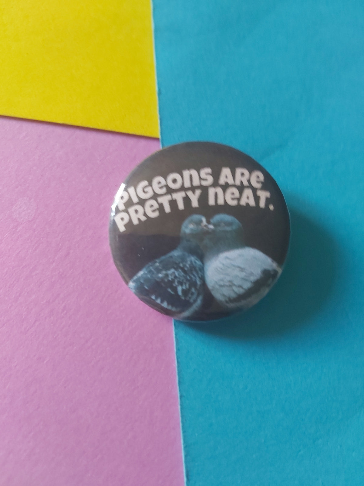 A round pinback button badge with a glossy finish, featuring the text 'Pigeons are pretty neat.' in white alongside image of two pigeons, displayed on a background of colored paper in blue, pink, and yellow.