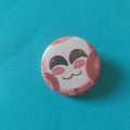 A round button badge featuring a stylised cutesy cartoon face with closed eyes on a white mask face and red ribbon hands, set against a solid teal-blue backdrop.