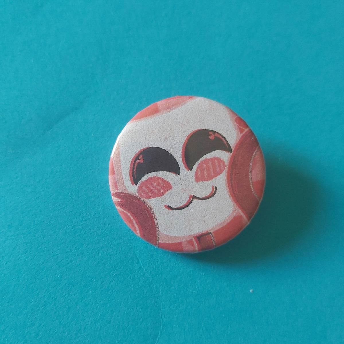 A round button badge featuring a stylised cutesy cartoon face with closed eyes on a white mask face and red ribbon hands, set against a solid teal-blue backdrop.