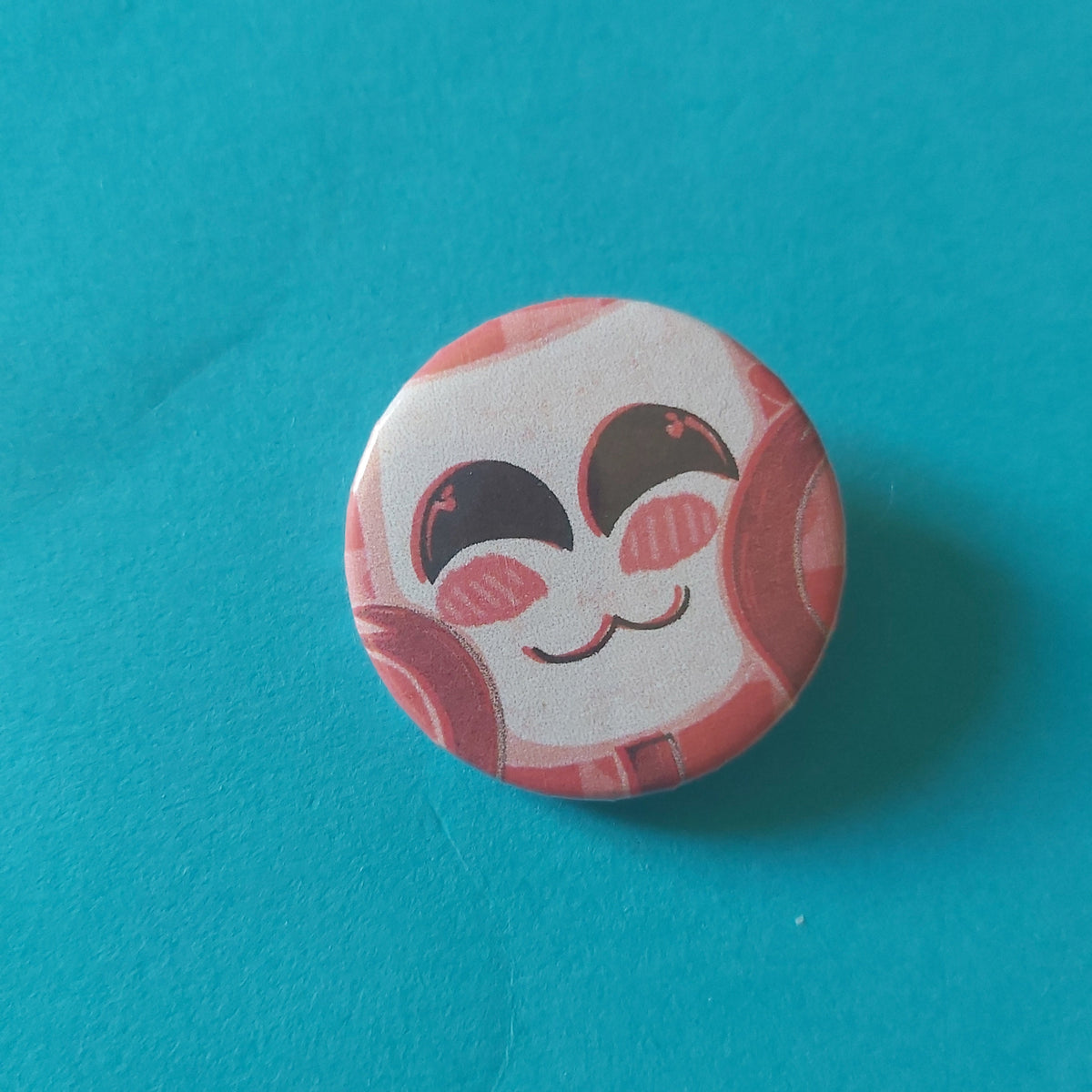 A round button badge featuring a cute cartoon face with pink closed eyes set against a solid turquoise background.