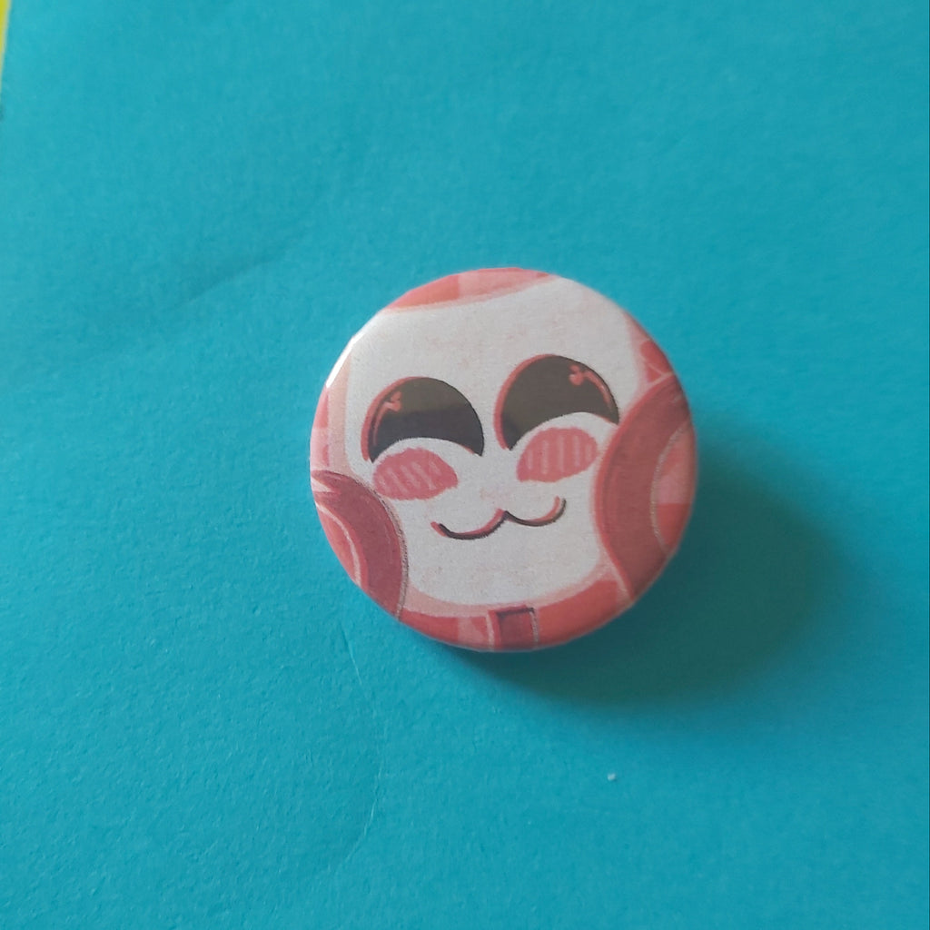 A round pinback button badge featuring a cute pink and white cartoon face, placed on a bright turquoise background with small yellow and pink paper corners visible.