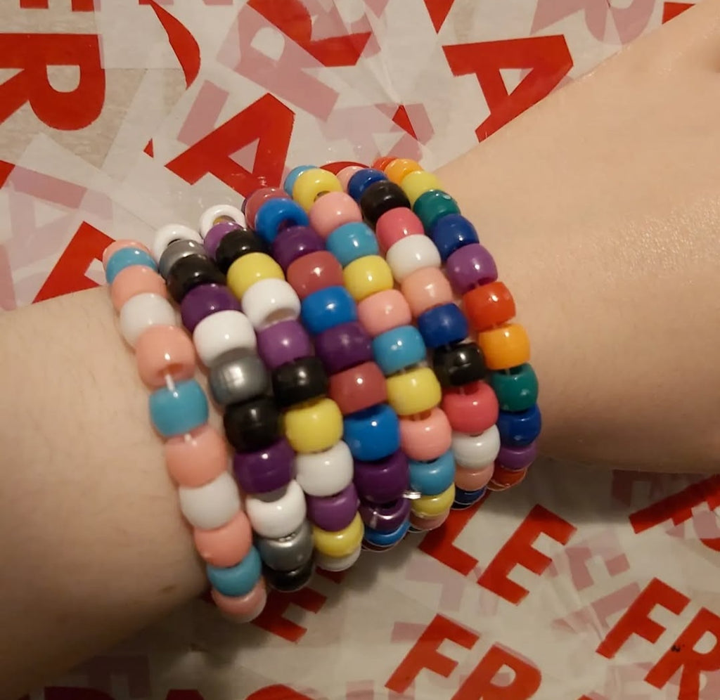 A close-up of a person's wrist wearing a stack of colorful beaded stretch bracelets. The bracelets feature multiple colors including pink, blue, yellow, purple, orange, black, white, and gray, arranged in a repeating pattern.