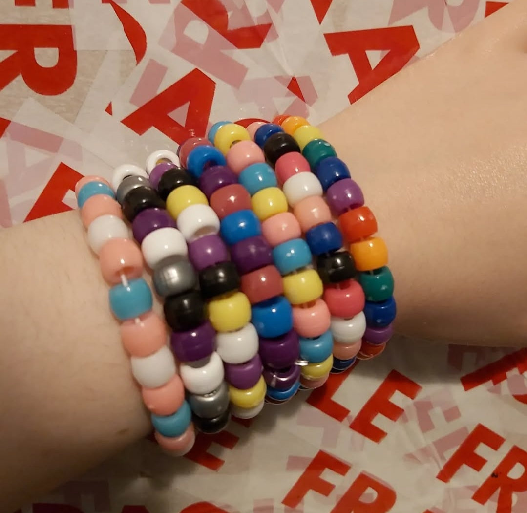 A close-up of a person's wrist wearing a stack of colorful beaded stretch bracelets. The bracelets feature multiple colors including pink, blue, yellow, purple, orange, black, white, and gray, arranged in a repeating pattern.