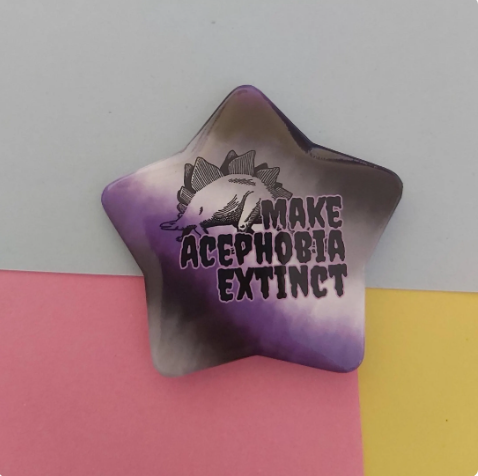 A star-shaped pin featuring a black line art illustration of a dinosaur and the text 'MAKE ACEPHOBIA EXTINCT' in black lettering. The pin has a purple, grey, white and black tie-dye gradient background.