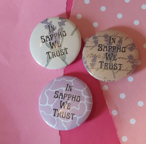Three round pin-back buttons with the phrase 'IN SAPPHO WE TRUST' arranged on pink paper. The top-left button is off-white with lavender flowers and dark text; the top-right button is beige with lavender flowers and script text; the bottom button is light purple with a marbled pattern and dark text.