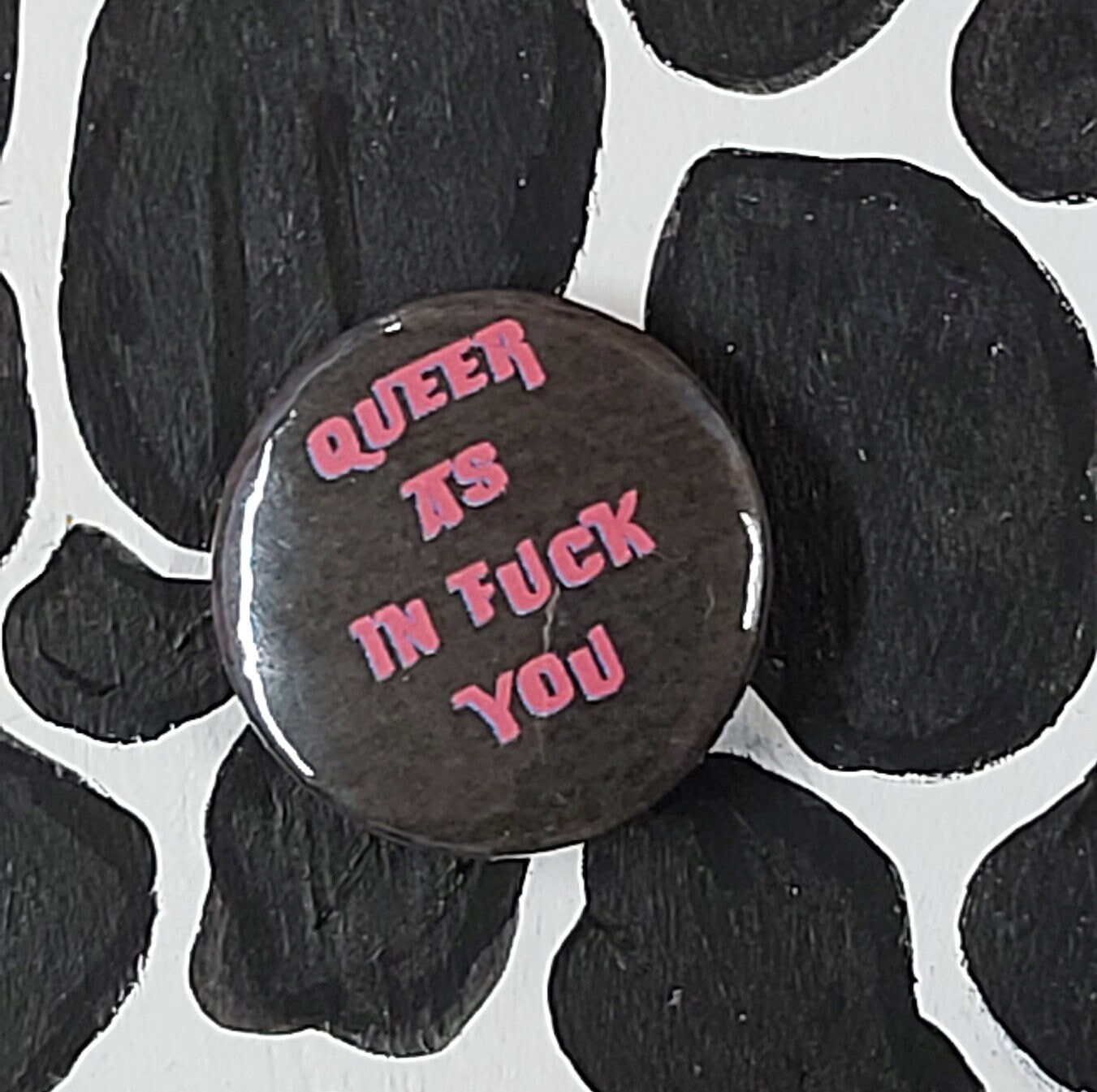 A round pinback button with the text 'QUEER AS IN FUCK YOU' in pink lettering on a black background, placed on a black and white cow-print surface.
