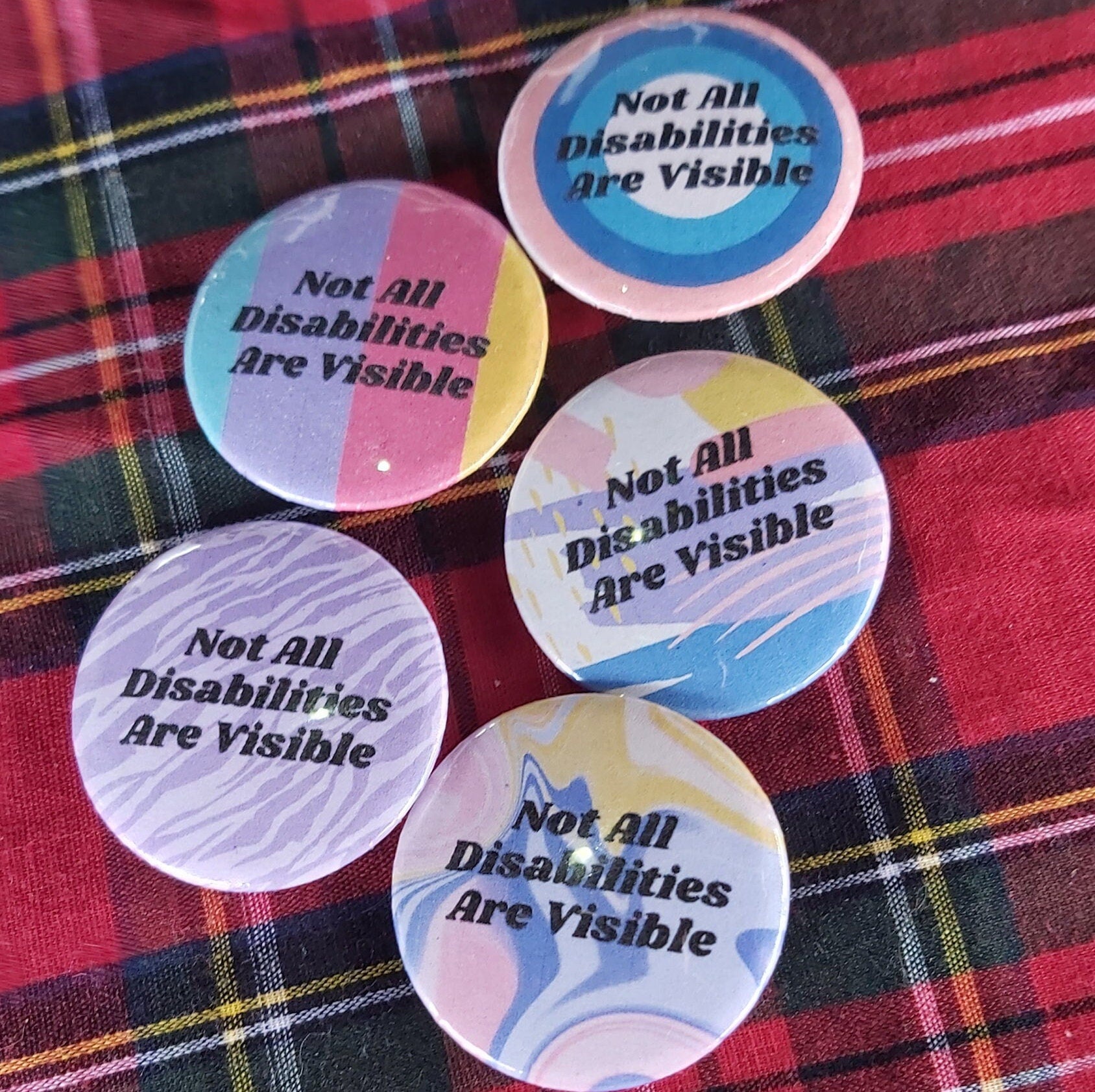 Five round button badges with the text 'Not All Disabilities Are Visible' in black, arranged on a red and yellow plaid fabric background.