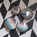 Four round glossy pinback buttons featuring the disability pride flags, displayed on a black-and-white geometric background.
