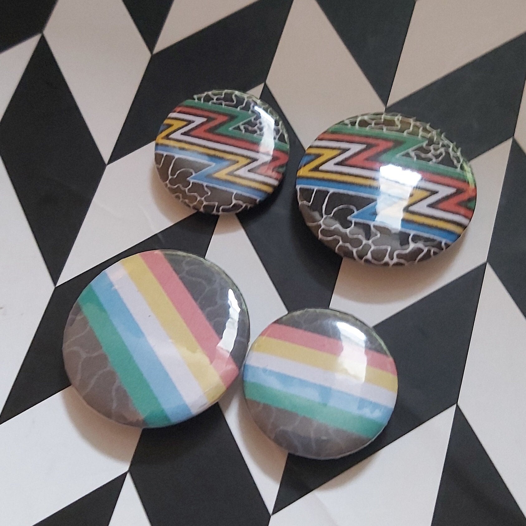 Four round glossy pinback buttons featuring the disability pride flags, displayed on a black-and-white geometric background.