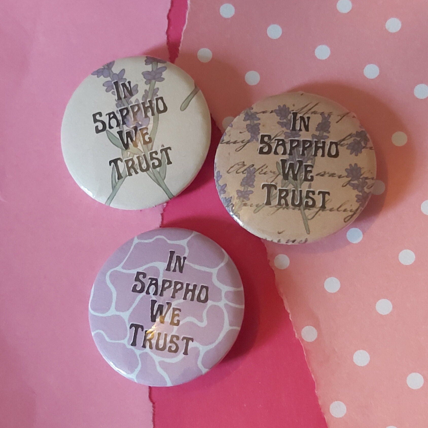 Three round pin-back buttons with the phrase 'IN SAPPHO WE TRUST' in dark lettering, arranged on a pink background with white polka dots. The buttons feature different decorative designs: one with lavender illustrations on a cream background, one with script and floral motifs on a tan background, and one with a purple and white abstract pattern.