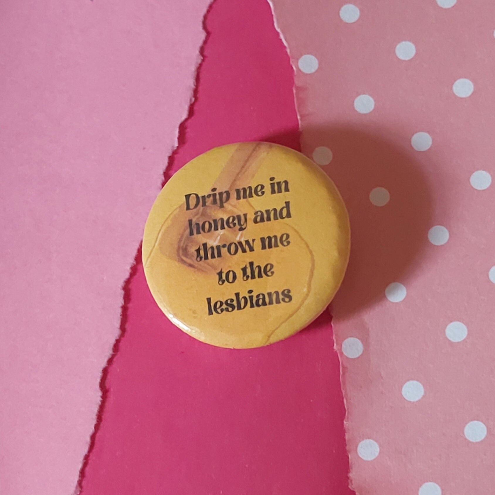 A round pinback button with black text reading 'Drip me in honey and throw me to the lesbians', placed on a pink background with torn paper and white polka dots.