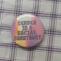 A round pinback button badge featuring the text 'GENDER IS A SOCIAL CONSTRUCT' in dark gray lettering over a pastel rainbow gradient background, placed on a purple and white plaid fabric surface.