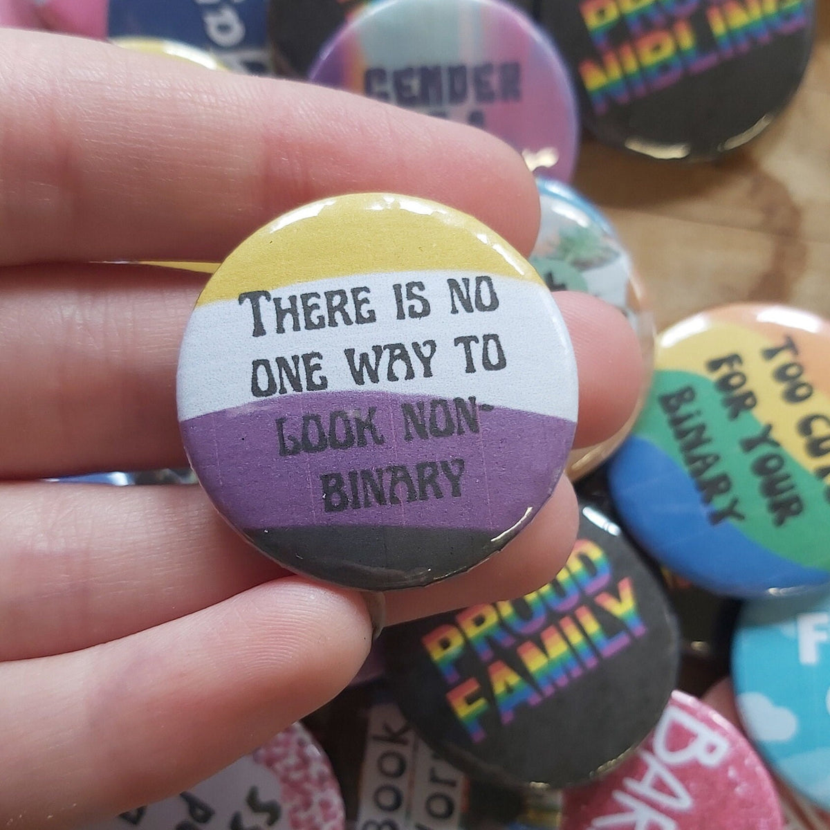 A hand holding a round pinback button with the text 'THERE IS NO ONE WAY TO LOOK NON-BINARY' over yellow, white, and purple stripes. Other colourful pride-themed buttons are scattered in the background.