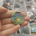 A hand holding a round pinback button featuring a frog photo and the word 'GAY' in white letters, with a background of a clear organiser filled with other buttons.