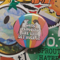 A round pinback button badge featuring a rainbow background and a penguin illustration with the text 'SOME PENGUINS ARE GAY, GET OVER IT.' placed on top of a colourful, sticker-covered surface.