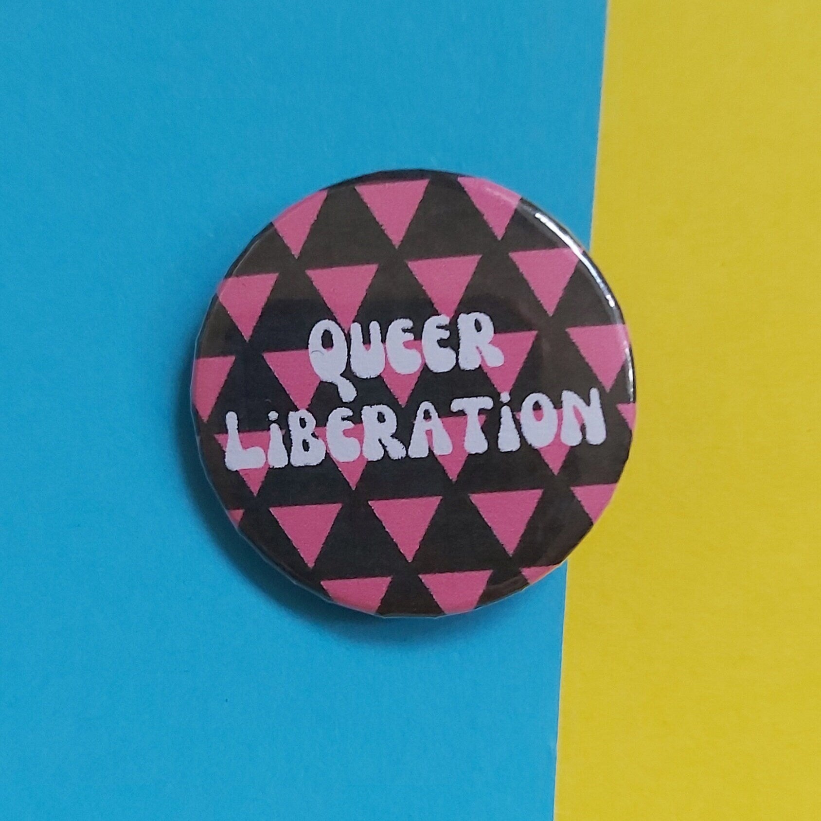 A round pinback button with a black background, pink geometric triangles pattern, and the words 'QUEER LIBERATION' in white. The button is photographed on a split blue and yellow background.