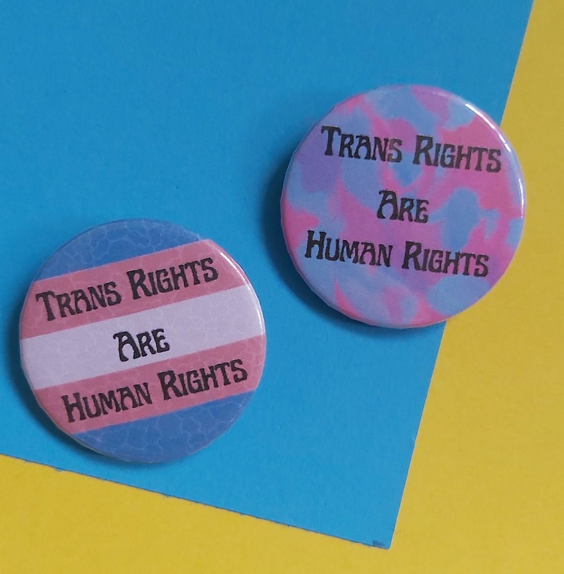 Two round pin-back buttons with the text 'TRANS RIGHTS ARE HUMAN RIGHTS' displayed on a split blue and yellow background.