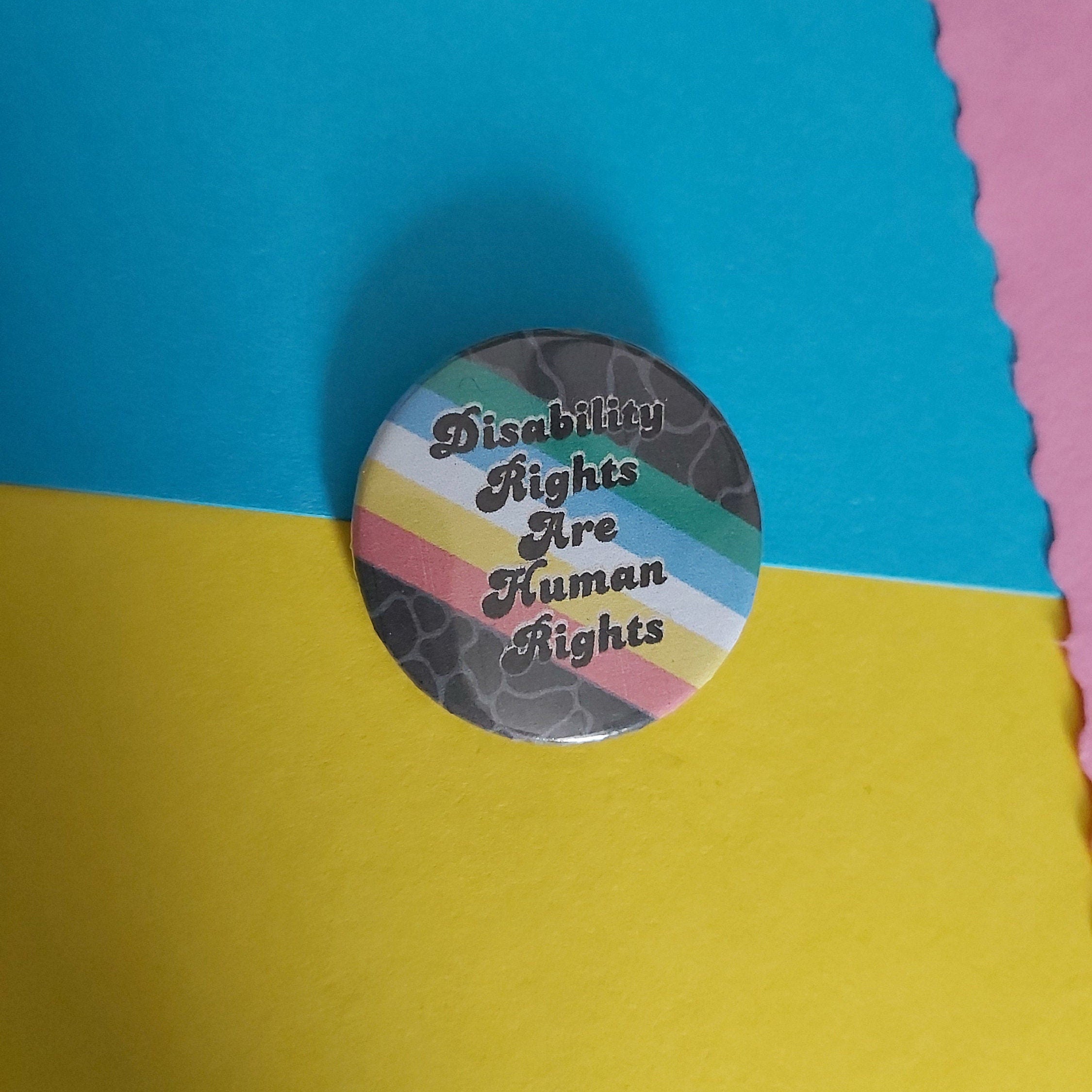 A round pinback button with the text 'Disability Rights Are Human Rights' in black script over a disability pride flag background, placed on a yellow and blue paper backdrop.