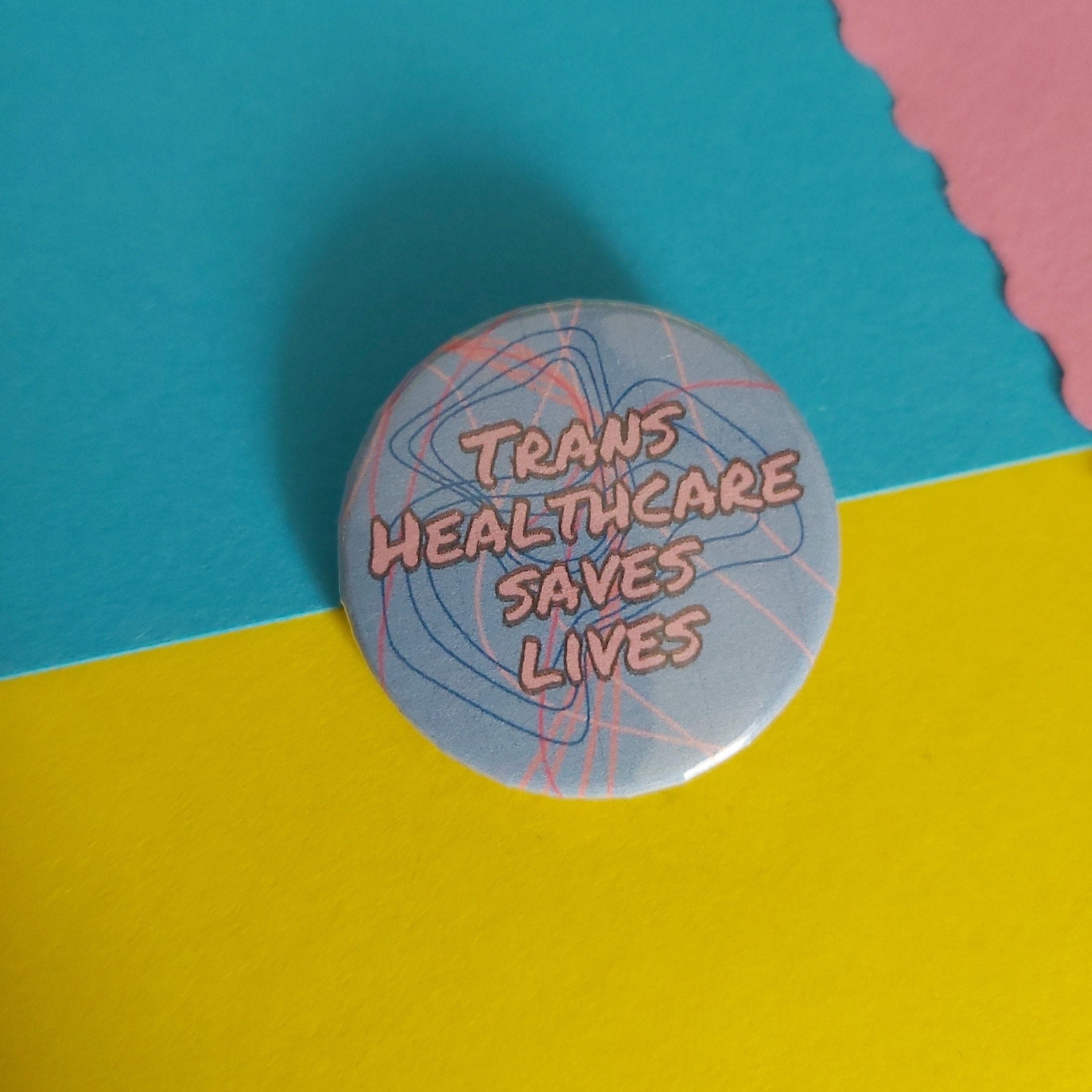 A round pinback button with the text 'TRANS HEALTHCARE SAVES LIVES' in pink lettering over a light blue background with abstract line art, set against a split yellow and turquoise paper backdrop.