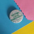A round pinback button badge with the text 'SUPPORT LOCAL BIODIVERSITY' in green, surrounded by light green nature illustrations, placed on a colorful background of blue, pink, and yellow paper.